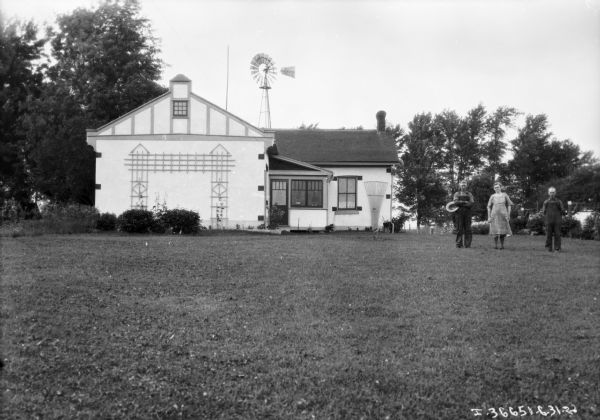 Two men and a woman are posing in the yard of a farmhouse. An Aermotor windmill and trees are behind the house.