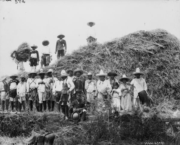 A large group of people, men, women, and children, are posing in a field near a large stack of hay.