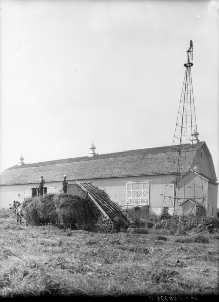 View across field towards men on a horse-drawn wagon using a hay loader fill the wagon with hay from the field. A windmill and a barn are in the background.