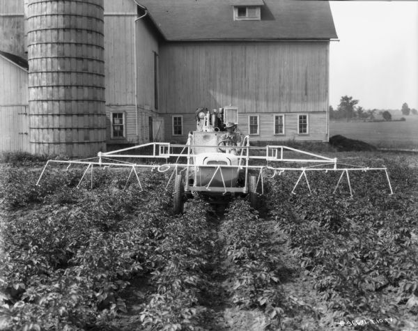 Front view of a man driving a tractor in a field with sprayers mounted on the front. In the background is a silo and a barn.