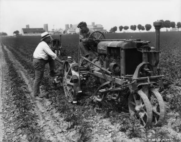 A man is standing with his foot on the cultivator attached to a tractor, talking to a man sitting on the McCormick-Deering Farmall tractor in a field. There is another tractor sitting in the field behind the men. Farm buildings are in the far background. (Perhaps at International Harvester model farm).