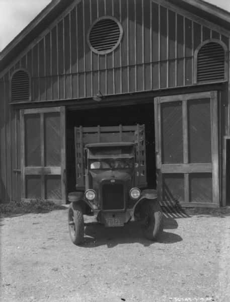 Two men are sitting in the front seat of a delivery truck, with stake body, exiting a barn.