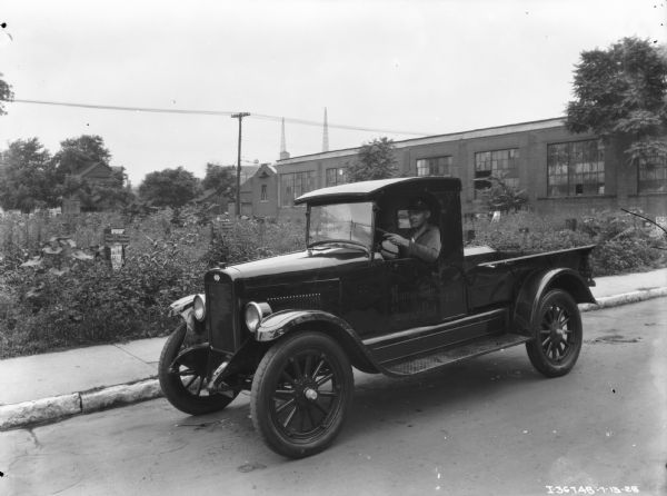 Three-quarter view from front of a man driving a truck down a street. The sign on the driver's side door reads: "Kansas City Journal Post." Two signs on posts among the plants in the background read: "Ralph T. Harding's Worm Medicine." There is a garden and a large building in the background.