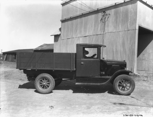 View towards the passenger side of a truck towards a woman sitting in the driver's seat. There is a tall, metal sided building behind the truck, along with other wooden buildings. The woman is wearing a brimmed hat with ball fringe.