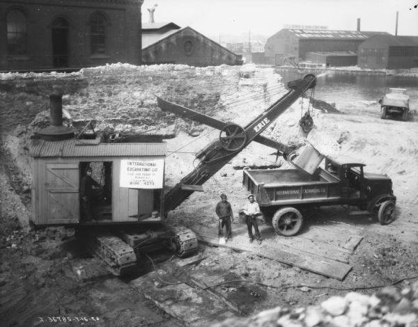 Elevated view of men standing near a steam shovel and International Excavating truck. There is a river in the background.