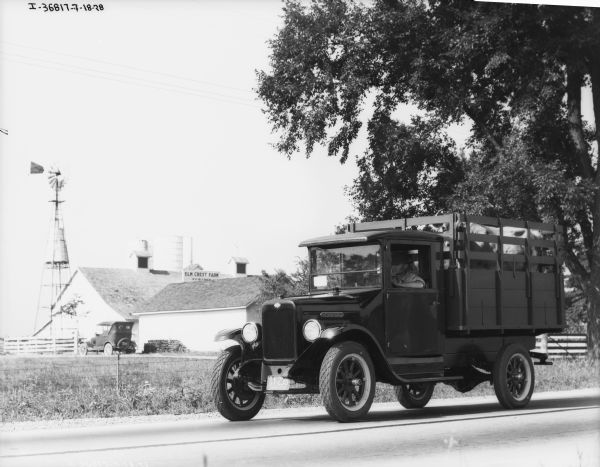 View of a man driving a livestock delivery truck on a road. In the background is the Elm Crest Farm.