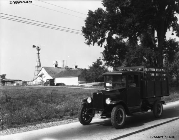 View of a man driving a livestock delivery truck on a road. In the background is the Elm Crest Farm.