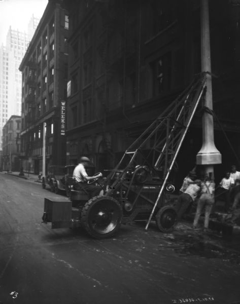View down street towards a man using a McCormick Deering hoist. A group of men are manually setting a lamppost or other object onto a sidewalk. 