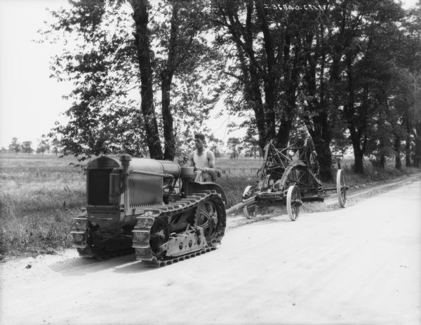 View across road towards a man driving a continuous track industrial tractor pulling a man on a road grader.