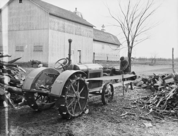 A man is using a McCormick-Deering 10-20 to belt drive a saw. The man is cutting wood, and there is a pile of wood on the right. In the background are barns.