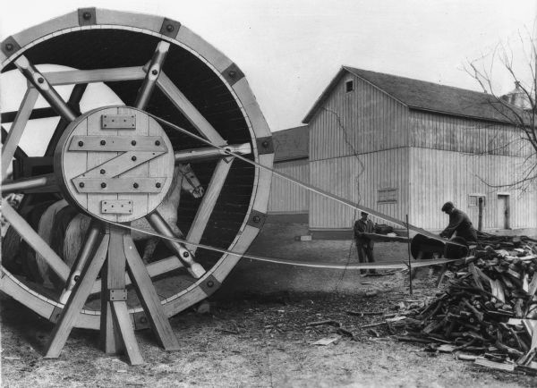 Graphic illustrating horse power. Horses are depicted powering a large wooden wheel to power a saw. Two men are working on the saw to cut up wood. Barns are in the background.
