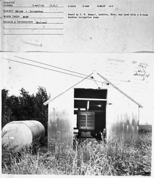 A UD-14A in a small building with an open door. There is a storage tank on the left. Subject: "UD-14A — Irrigation." Where Taken: "MidW." Information with photograph reads: "Owned by J. T. Rempel, Lushton, Nebr. and used with a 8-inch turbine irrigation pump."