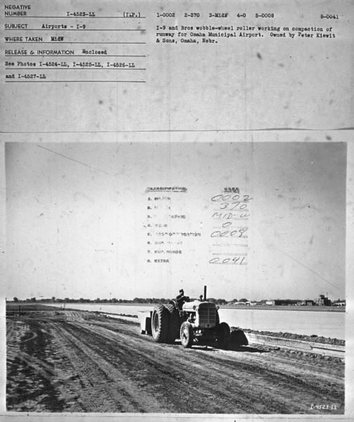 View towards a man driving a tractor to compact a runway. Subject: "Airports — I-9." Where Taken: "MidW." Information with photograph reads: "I-9 and Bros wobble-wheel roller working on compaction of runway for Omaha Municipal Airport. Owned by Peter Kiewit & Sons, Omaha, Neb."