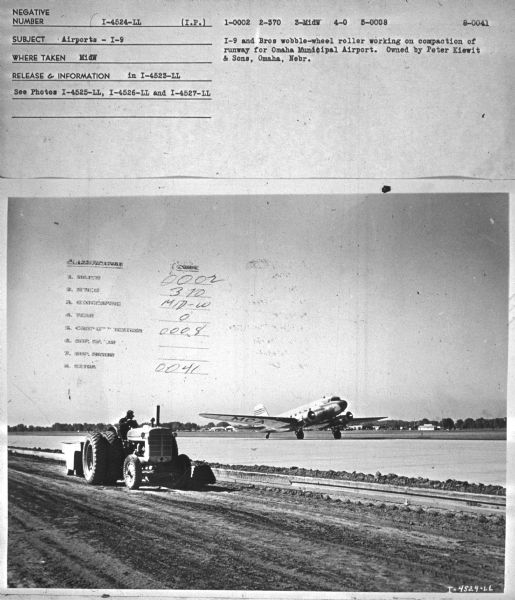 View towards a man driving a tractor to compact a runway. There is an airplane on the runway in the background. Subject: "Airports — I-9." Where Taken: "MidW." Information with photograph reads: "I-9 and Bros wobble-wheel roller working on compaction of runway for Omaha Municipal Airport. Owned by Peter Kiewit & Sons, Omaha, Neb."