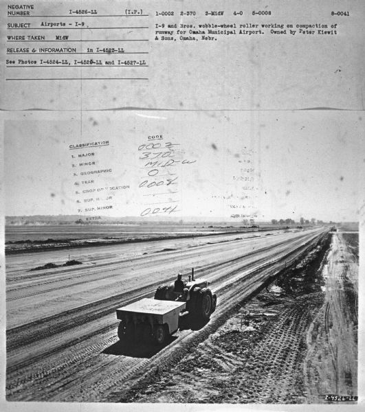 Elevated view of a man driving a tractor to compact a runway. Subject: "Airports — I-9." Where Taken: "MidW." Information with photograph reads: "I-9 and Bros wobble-wheel roller working on compaction of runway for Omaha Municipal Airport. Owned by Peter Kiewit & Sons, Omaha, Neb."