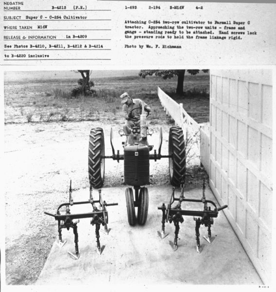 Slightly elevated view from front of a man sitting on a Farmall Super C tractor. Subject: "Super C — C-254 Cultivator." Where Taken: "MidW." Information with photograph reads: "Attaching C-254 two-row cultivator to Farmall Super C tractor. Approaching the two-row units — frame and gangs — standing ready to be attached. Hand screws lock the pressure rods to hold the frame linkage rigid. Photo by Wm. F. Eichmann."	