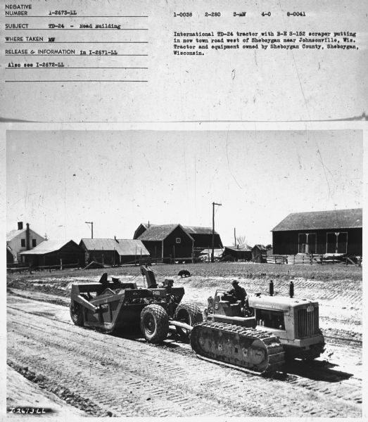 View towards a man working on building a road. In the background is a dog, and farm buildings. Subject: "TD-24 — Road Building." Where Taken: "MW." Information with photograph reads: "International TD-24 Tractor with B-E S-152 scraper putting in new town road west of Sheboygan near Johnsonville, Wis. Tractor and equipment owned by Sheboygan County, Sheboygan, Wisconsin."