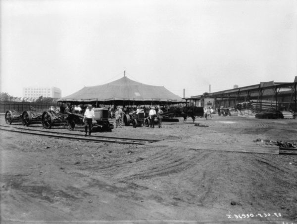 View across railroad tracks towards IH implements near a large, circular tent. People are walking on the grounds, and more people are inside the tent. A large fence and buildings are in the background.