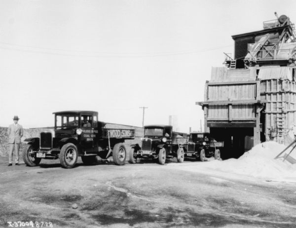 A man is standing near a row of trucks near an industrial building. Signs on the truck read: "Buildings Supplies, Coal, Coke, Brick."