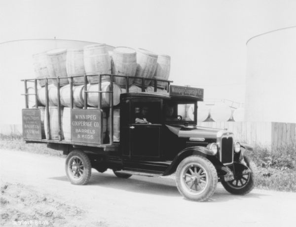 Men are sitting in the cab of a truck. The back is loaded with barrels.