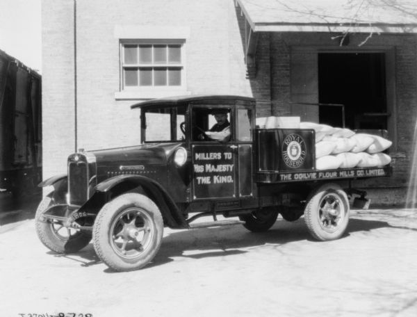 A man is sitting in the driver's seat of a delivery truck. The back is piled with sacks of flour. The signs on the truck read: "Millers to His Majesty the King. Royal Household. The Ogilvie Flour Mills Co., Limited."