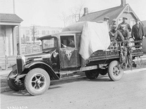 A man is sitting in the driver's seat of a truck. A group of men are sitting and standing in the back of the truck, which has a stake body. A cloth is covering the back of the truck just behind the cab. Buildings are in the background.
