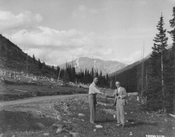 Two men in business suits shaking hands while standing near a road. There is a cleared forest behind them, and a mountain peak is in the distance.