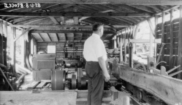 A man is standing in a sawmill. An engine in the background on the left is powering the sawmill.