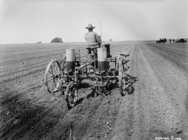 Rear view of a man using a Farmall tractor in a field. In the background on the right are men standing near two automobiles.