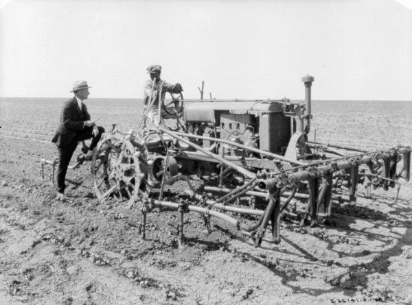A man wearing a suit is standing next to a man driving a Farmall tractor in a field. A cultivator is attached to the tractor.
