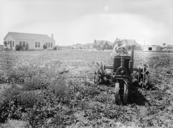 A man is using a Farmall tractor in a field to pull an implement. There are houses in the background.