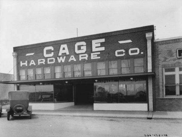 View from street towards the facade of a hardware store and dealership. The name on the store reads: "Cage." There are large show windows flanking the entrance, and automobiles are parked at the curb in front.