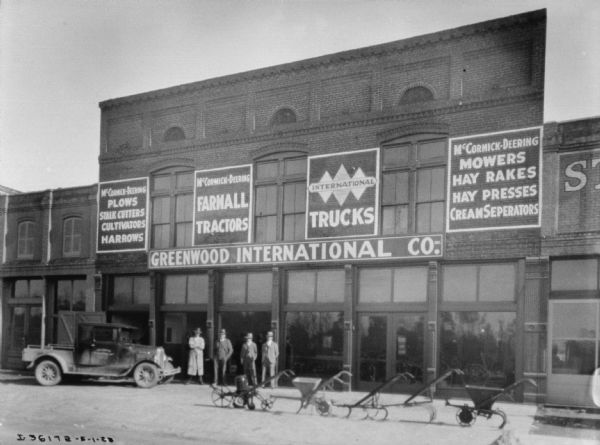 View from street towards storefronts. A group of men are standing on the sidewalk in front of the Greenwood International Co. There is a truck parked at the curb on the left. Agricultural implements are on display and lined up in the street.