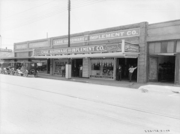 View from street towards the facade of a hardware store and dealership. Men are standing at the entrance. The name on the store reads: "Cage Hardware & Implement Co." There are large show windows flanking the entrance, and automobiles are parked at the curb in front.