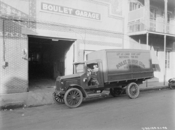 View across street towards a man sitting in the driver's seat of a truck. A sign on the truck reads: "Boulet Transp. Co. Inc. The truck is parked in front of a large building with a sign that reads: "Boulet Garage."