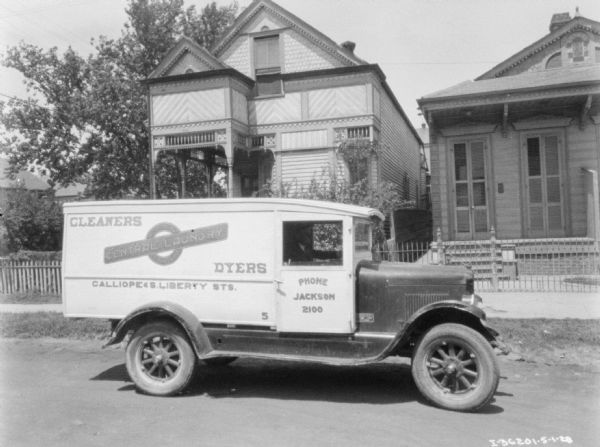 View across street towards a man sitting in the driver's seat of a truck parked in a neighborhood. The sign on the truck reads: "Central Laundry, Cleaners, Dyers."