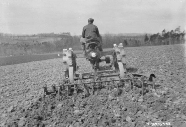 Rear view of a man driving a Farmall tractor to pull a cultivator.