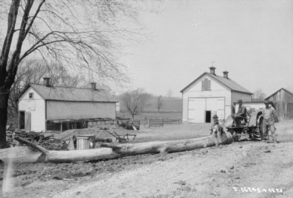 View of three men and a dog posing with a tractor. There are farm buildings in the background. A large tree trunk is attached to the back of the tractor with a chain.