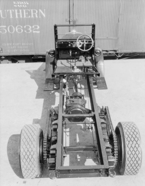View of an exposed chassis of a truck, which is parked outdoors near a railroad car. The truck has a hood and a windshield, and there is no driver's seat. The steering wheel is on the right side.