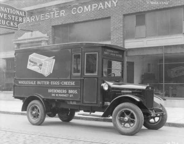 View across street towards the passenger side of a dairy delivery truck. The sign painted on the truck reads: "Bredenberg Bros., Wholesale Butter • Eggs • Cheese." The brick building behind the truck has a sign for the International Harvester Company.