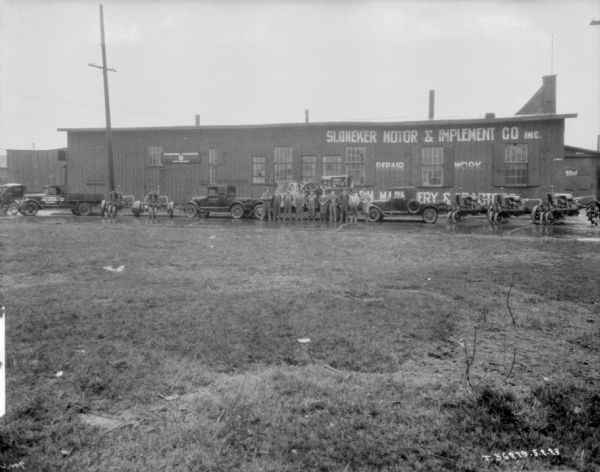 View across grassy field towards a group of people posing outdoors in the rain in front of Sloneker Motor & Implement Co. Inc.