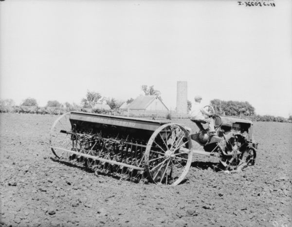 Three-quarter view from right rear of a young boy driving a Farmall tractor pulling a McCormick-Deering grain drill. Farm buildings and a silo are in the background.