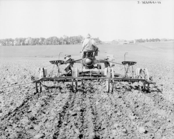 View from rear of a man using a Farmall tractor to pull a planter in a field. Farm buildings and trees are in the distance.