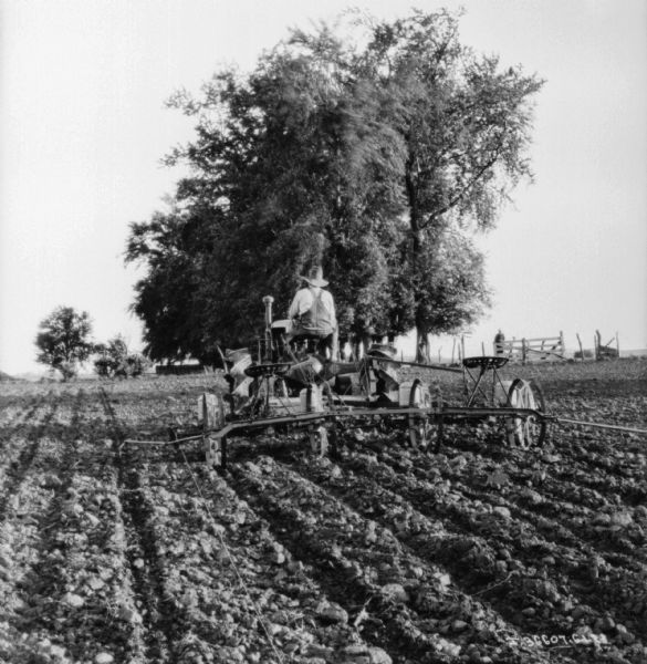 Rear view of a man using a Farmall tractor to pull a planter in a field. A man is standing near a gate in a fence in the background on the right.
