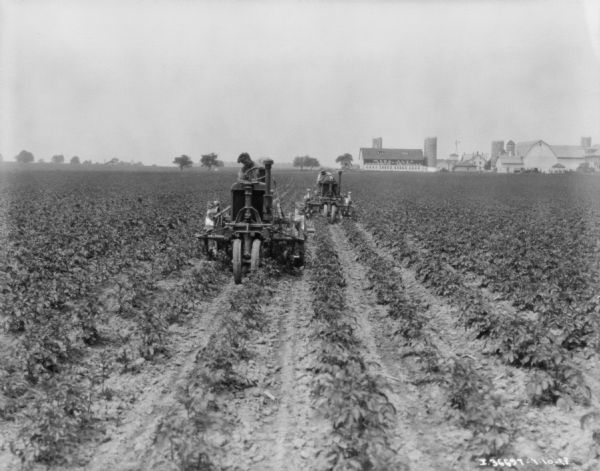 View towards two men driving Farmall tractors with cultivators in a field. There are farm buildings in the background.