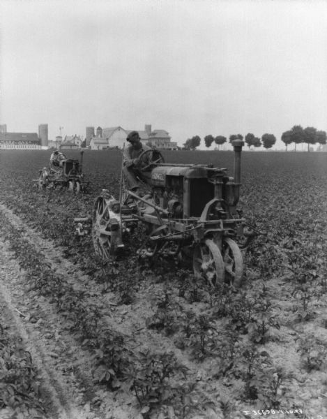 View towards two men driving Farmall tractors with cultivators in a field. There are farm buildings in the background.