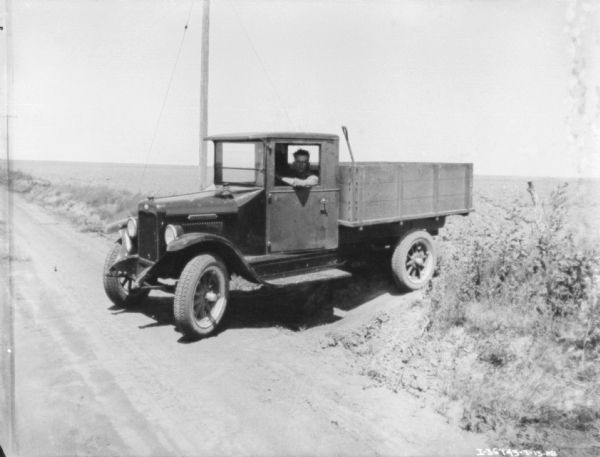 View down dirt road towards a man sitting in the driver's seat of a truck. There is a shovel or other implement in the wood-sided truck bed. The truck is backed up to the side of the road, with a field in the background.