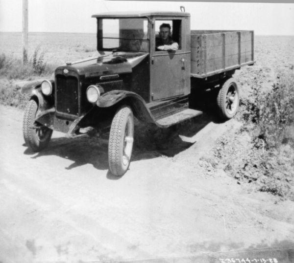 View towards a man sitting in the driver's seat of a truck. There is a shovel or other implement in the wood-sided truck bed. The truck is backed up to the side of the dirt road, with a field in the background.