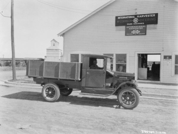 View of a man driving a truck on a road. There is a sign on a building behind the truck that reads: "International Harvester McCormick-Deering Farm Machines."