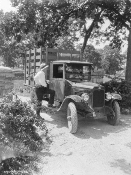 View from road towards a man standing near the passenger side of a truck while talking to the man in the driver's seat. The truck bed has a high stake body for livestock, and is parked at a gate.